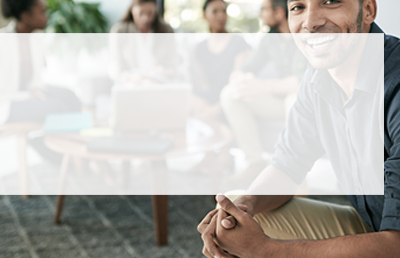 A man smiles towards the camera while a group of team members work in the background around a table.