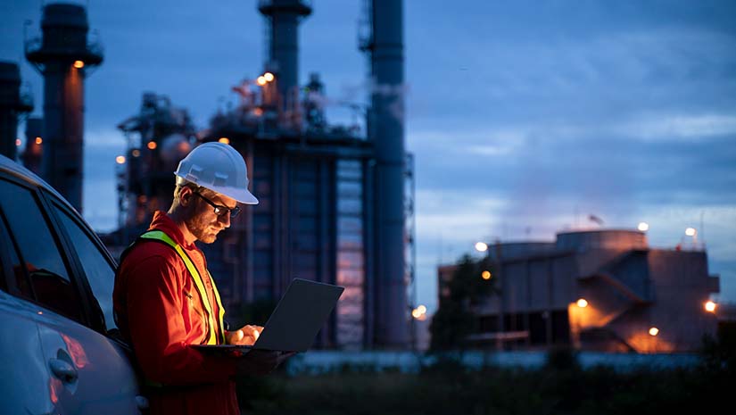 A side view of an young engineer wearing a helmet and using a laptop and hands free device to discuss a prolem with collegues during his night shirt in the oil rafinery.