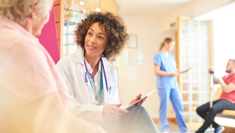 A female doctor sits in doctor’s waiting room with female patient taking notes and updating her medical records.