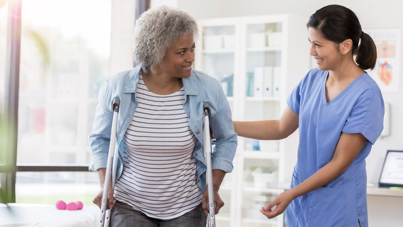 A senior woman stands on a pair of crutches in her physical therapist's office.