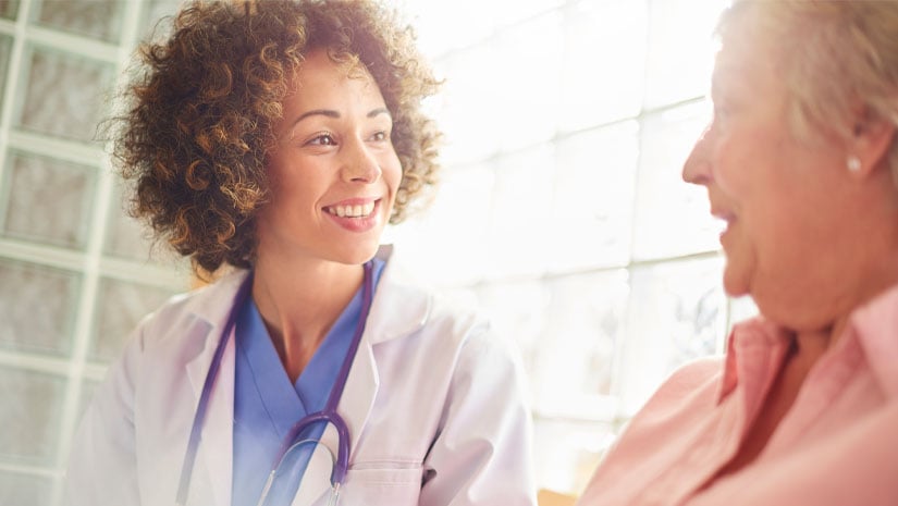 A female nurse sits in doctor’s waiting room with senior female patient taking notes and updating her medical records