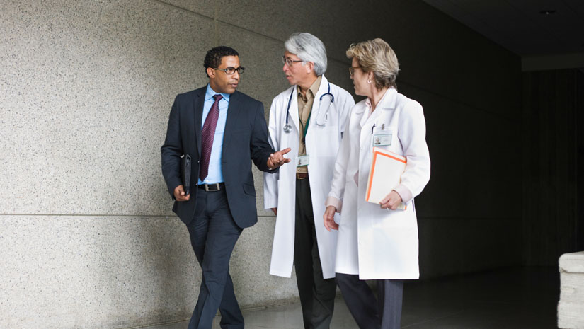 Two doctors speaking to businessman while walking down hallway.