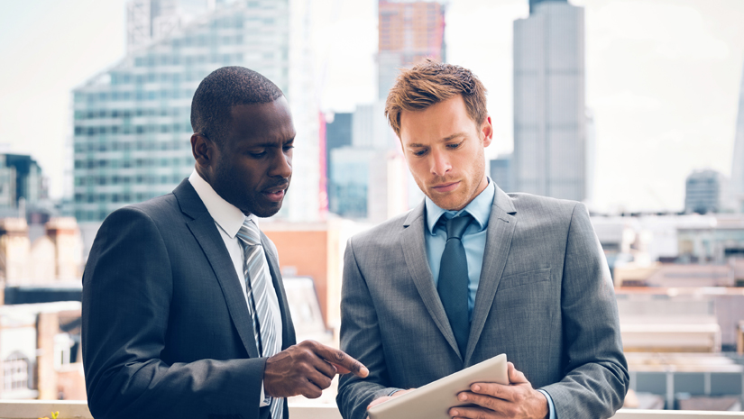 Two male realtors talking outside while pointing at a tablet.